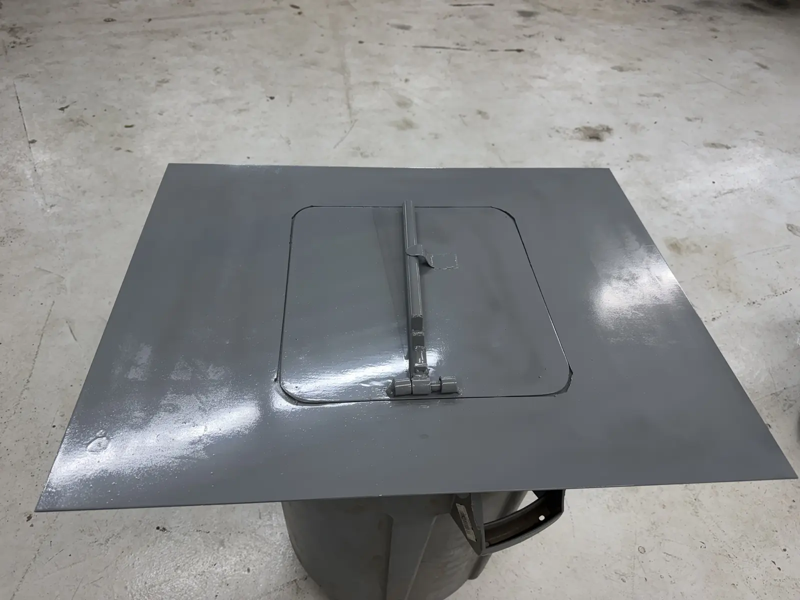 Gray painted sheet metal vent plate with a hinged flap and pipe lever, closed, resting on a trash can in the IronMann fabrication shop in Flint, MI
