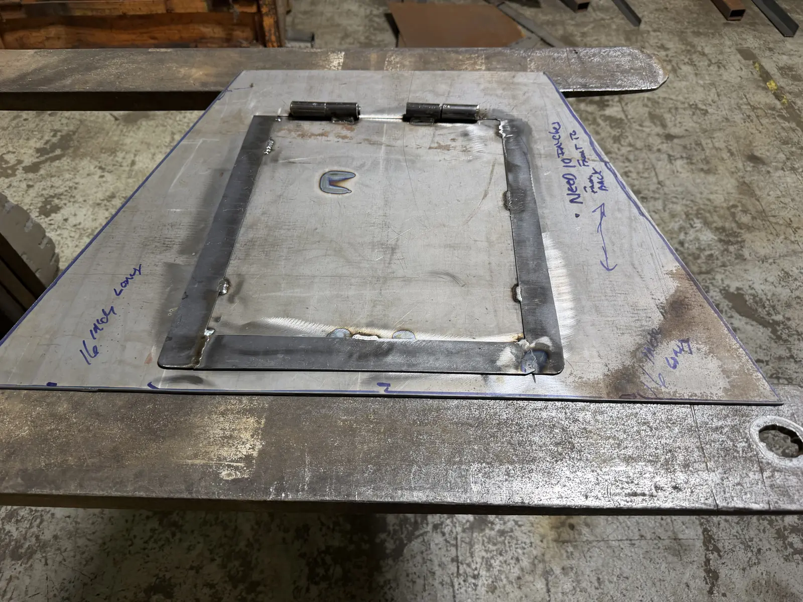 Bare steel vent plate on a welding table with a hinged flap, welded hinges and marker layout notes on the sheet