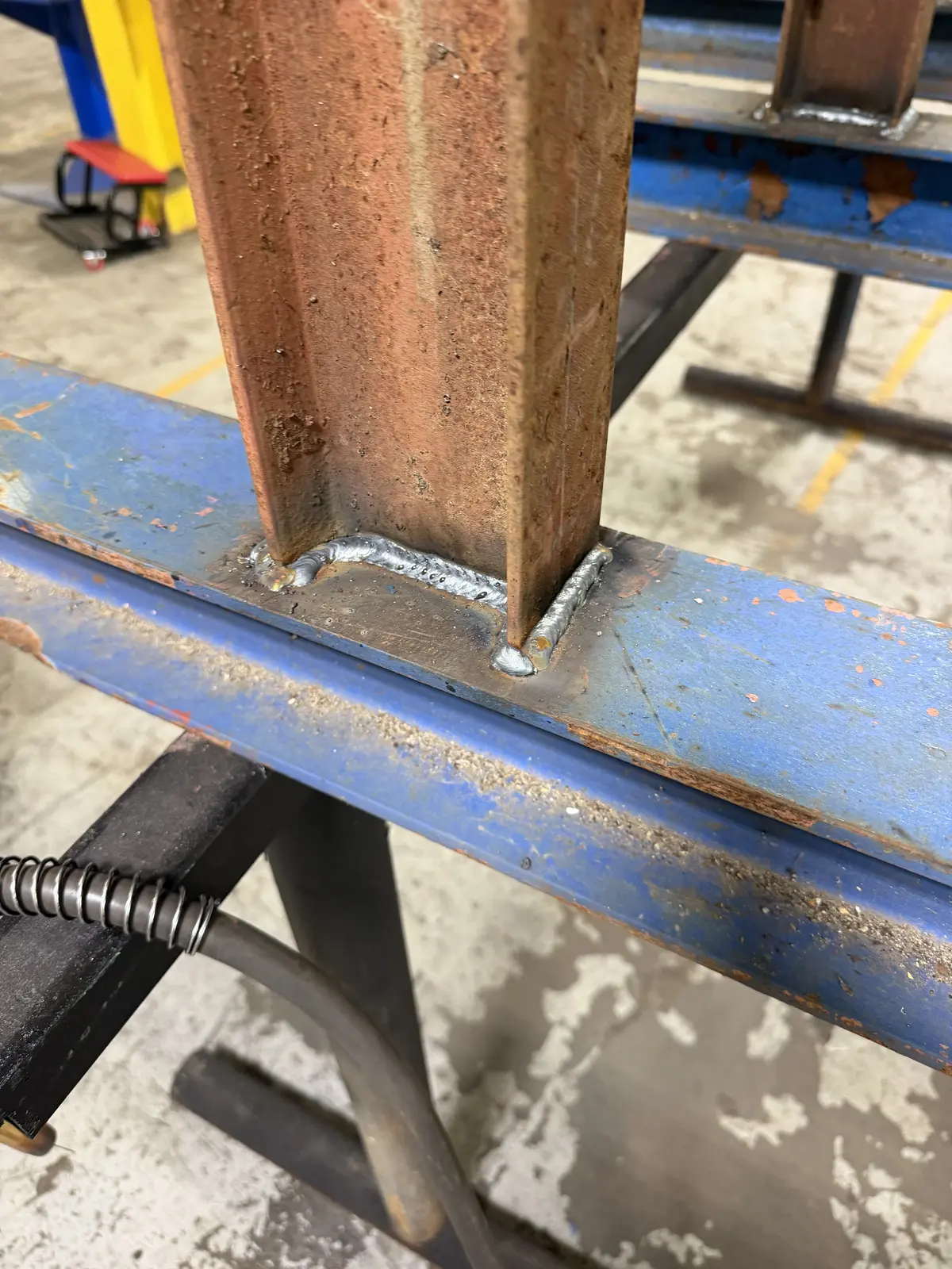 Close-up of the weld bead joining a rust-colored steel arm to a blue column on a cantilever rack built in Flint, MI
